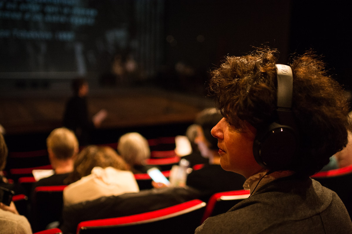 Dans une salle de théâtre aux fauteuils de velours rouges, une femme de dos porte un casque audio sur ses cheveux frisés.