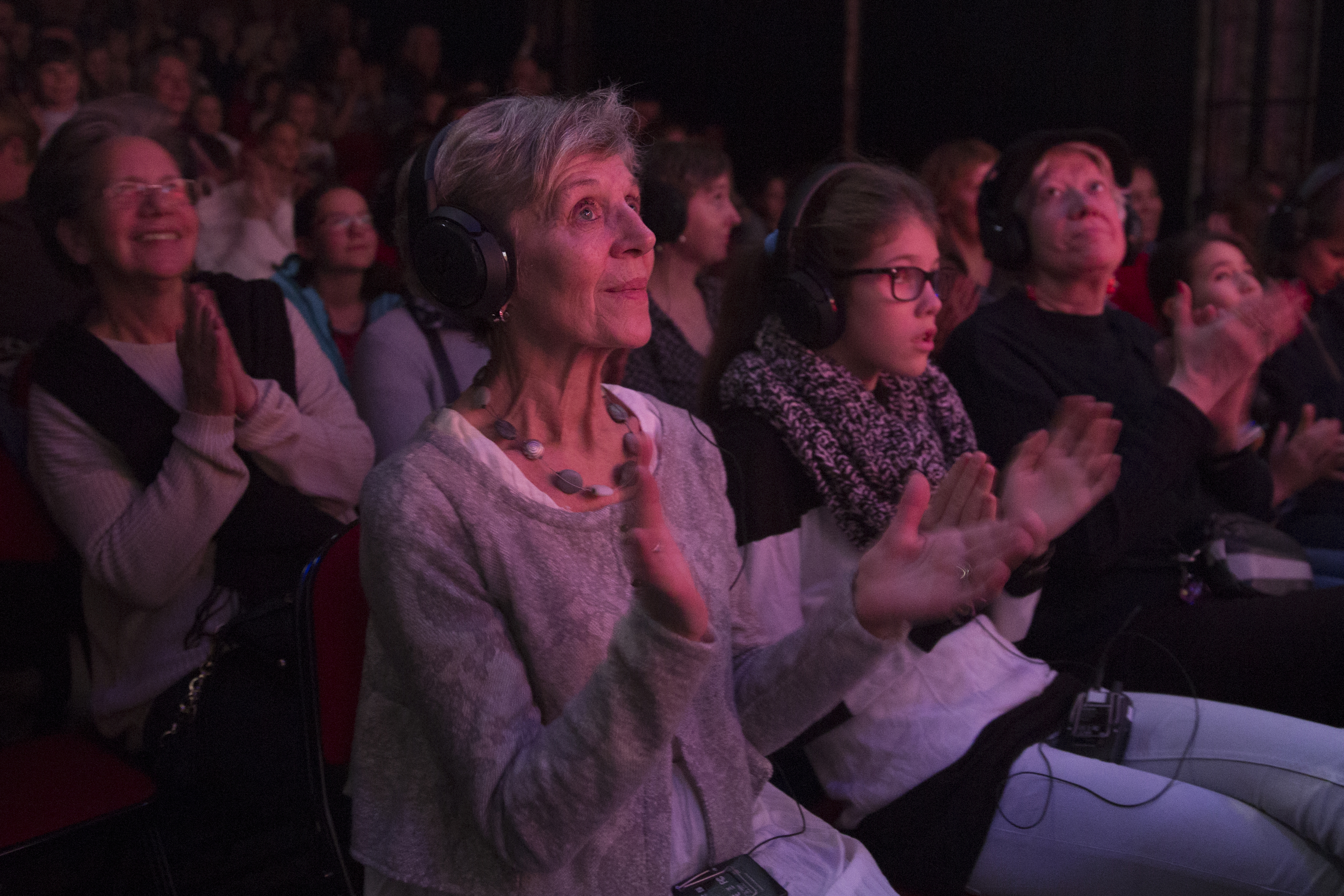 Descriptif de l'image: Une salle de théâtre bondée dans laquelle on voit une femme au premier rang qui applaudit. Elle porte un casque sur les oreilles. A ses côtés et derrière elles plusieurs personnes portent aussi des casques.