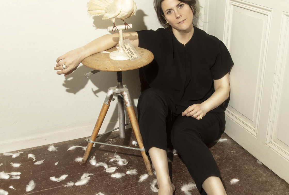 Une femme assise à même le sol, contre une  porte. Elle est vêtue d'un tshirt et pantalon noir, es genoux sont repliés. L'un de ses bras est posé sur un tabouret en bois sur lequel on devine une colombe empaillée. L'autre bras repose sur son genou. Son visage exprime une forme de lassitude. Le sol est jonché de quelques plumes.