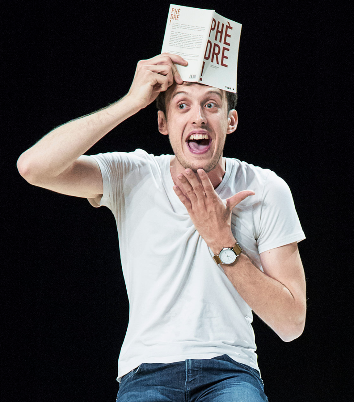 Portrait en plan américain d'un jeune homme vêtu d'un t-shirt blanc. De sa main droite il tient un livre sur son front. Le titre de ce livre est Phèdre. Sa main gauche est posée sous son menton. Il semble réjoui.