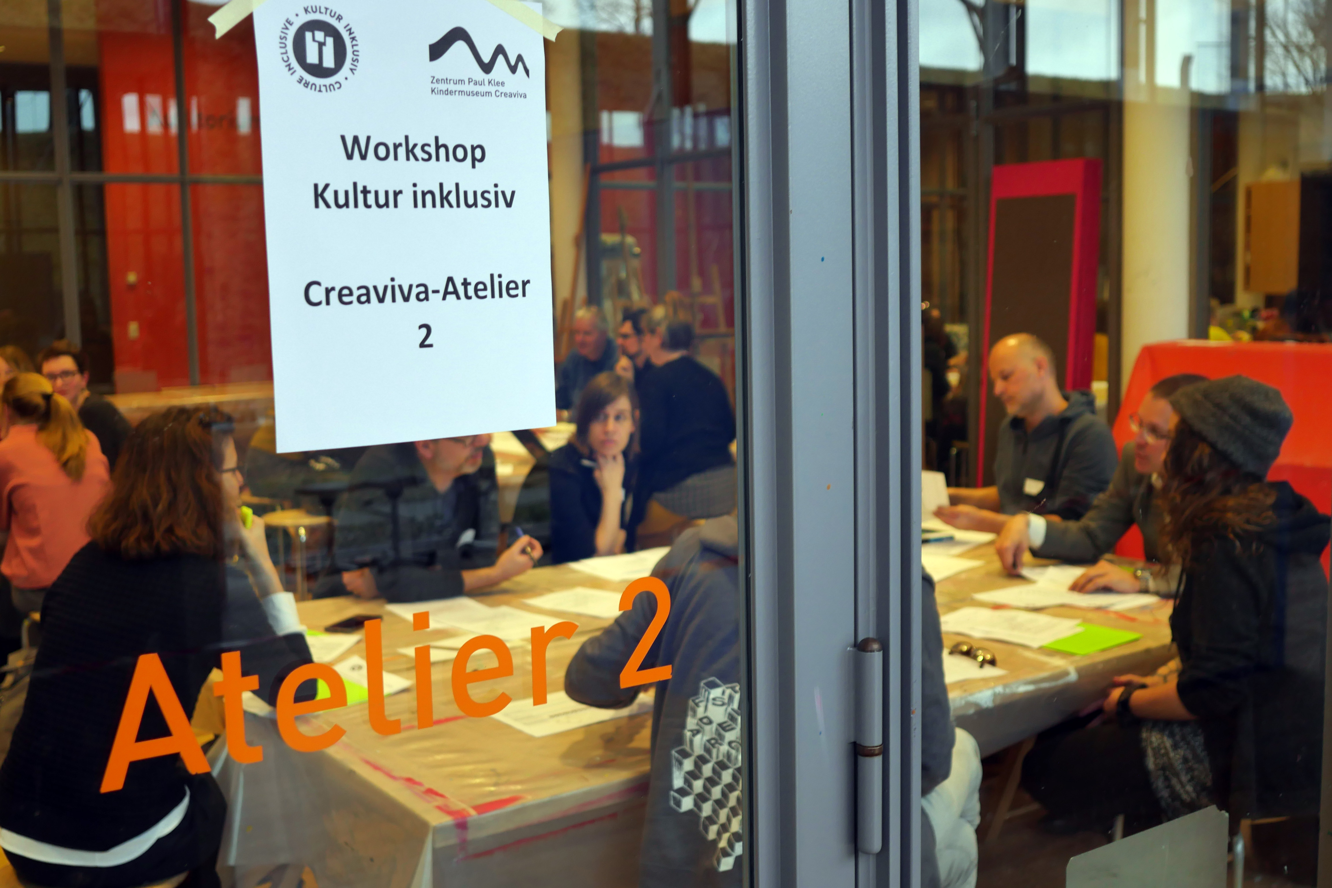 La photo représente l'atelier participatif. Derrière une porte transparente indiquant Atelier 2, on distingue deux grandes tables à dessin autour desquelles plusieurs personnes discutent.