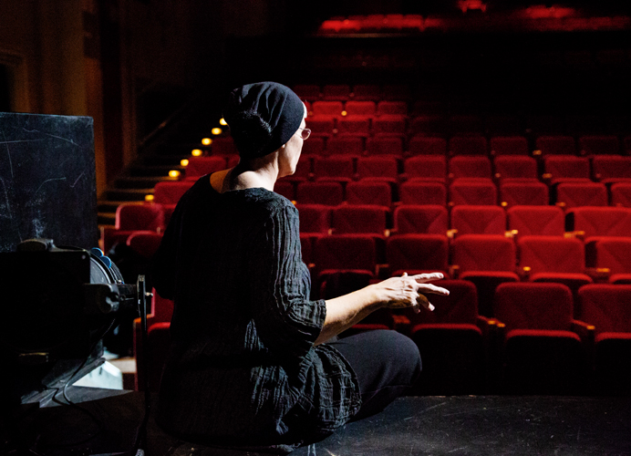 Description de la photo: Sur la photo une femme, de dos, vêtue de noir et portant un bonnet noir, est assise sur le bord d'une scène. Devant elle, une salle de théâtre vide composée de sièges rouges. Ce moment correspond au réglage de la lumière sur l'interprète, réalisé avec les techniciens lumière du théâtre.