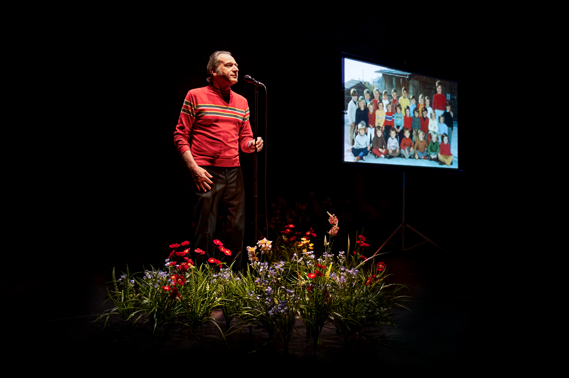 Une photo de classe est projetée sur un écran blan. Le comédien positionné derrière un parterre de fleurs des champs est vêtu d'un pull rouge et se tien debout face à un micro sur pied