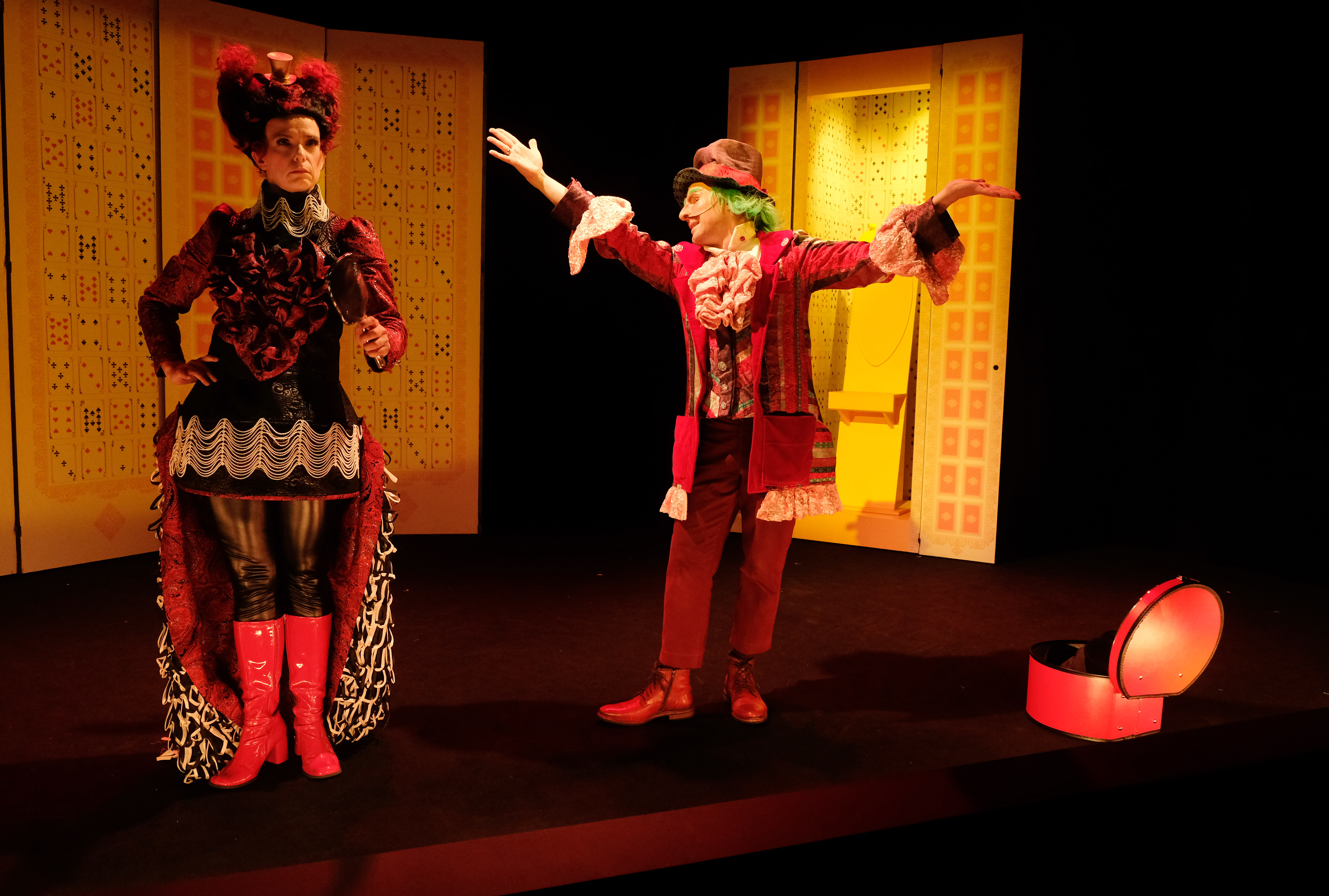 Descriptif de l'affiche du spectacle: Sur une scène de théâtre, un personnage clownesque lève les bras au ciel de manière grandiloquente, il parle à une femme portant une perruque et des bottes rouges.