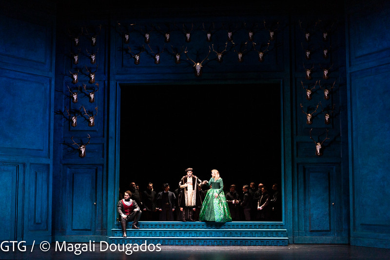 Décor représentant l'intérieur d'un palais bleu. Au centre un large escalier composé de trois marches. Un homme à l'allure de noble est assis sur l'une d'elle, à sa gauche un homme avec une tunique blanche, un long manteau et un chapeau tient le bras d'une femme vêtue d'une robe de bal verte.