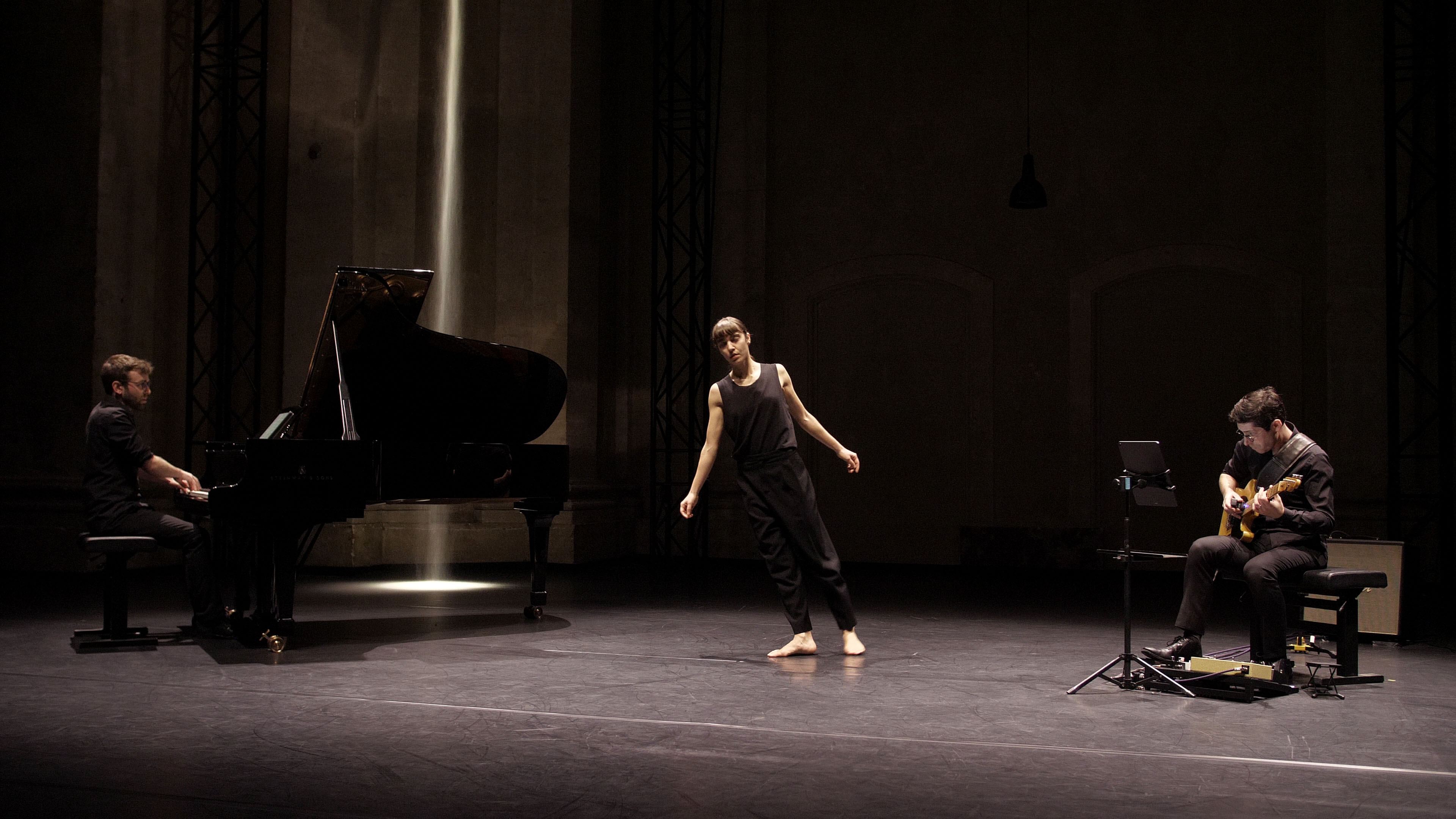 Sur scène à gauche un pianiste devant un piano à queue, au centre une danseuse dont le corps tangue vers le piano, à droite un musicien assis qui joue de la guitare