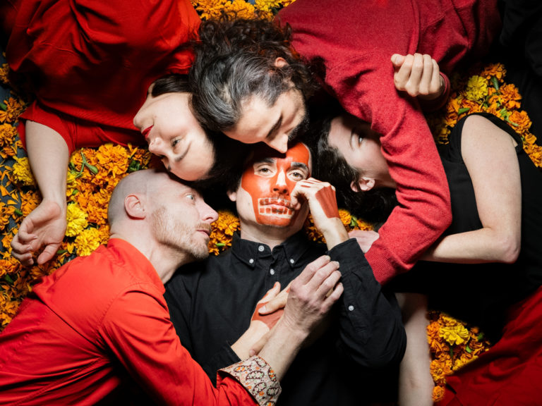 Cinq personnes sont couchées sur un tapis de fleurs oranges (tagetes). Elles sont placées en étoile, collées tête contre tête. Seule une personne regarde l'objectif. Il s'agit d'un homme vêtu d'une chemise noire et dont le visage est grimé en tête de mort.