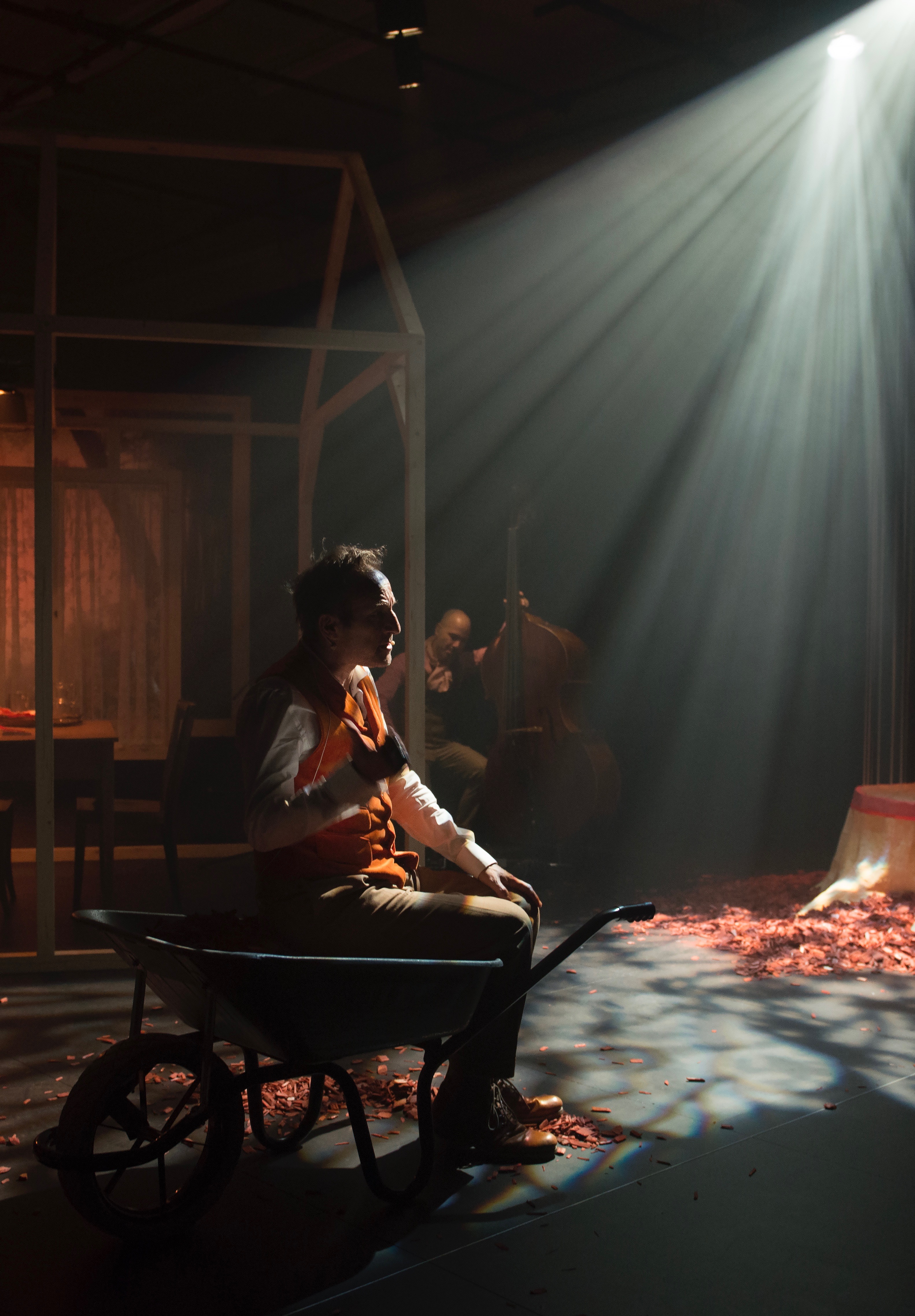 Sur une scène de théâtre un homme de profil est assis dans une brouette. Il porte un gilet orange. Une lumière vive éclaire le plateau devant lui.