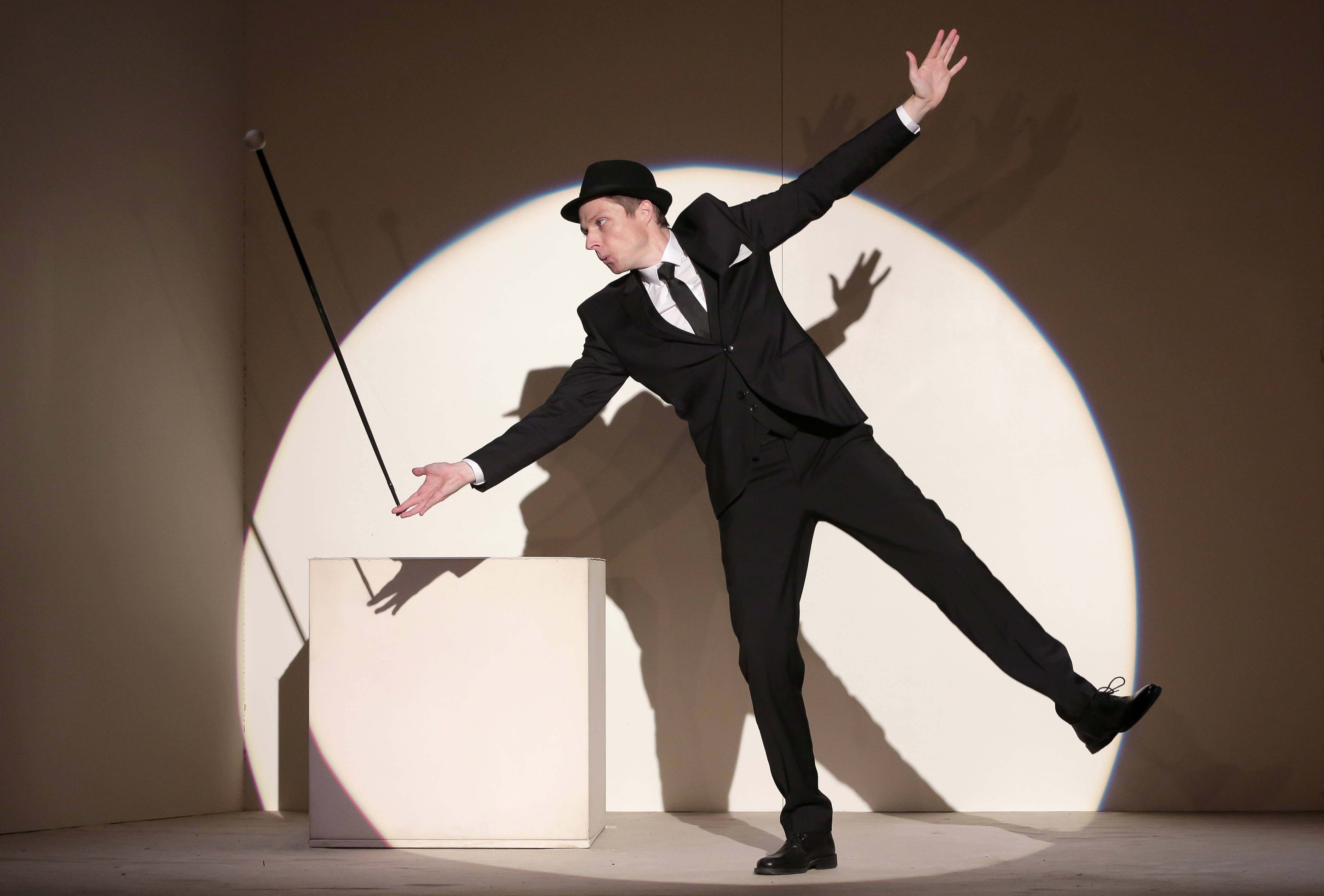 Un homme vêtu d'un costard noir, chemise blanche, cravate et chapeau noir est en équilibre sur une jambe, penché sur le coté bras écartés. Sur le bout de ses doigts une canne tient en équilibre. Il se tient au milieu d'un cercle de lumière blanche.
