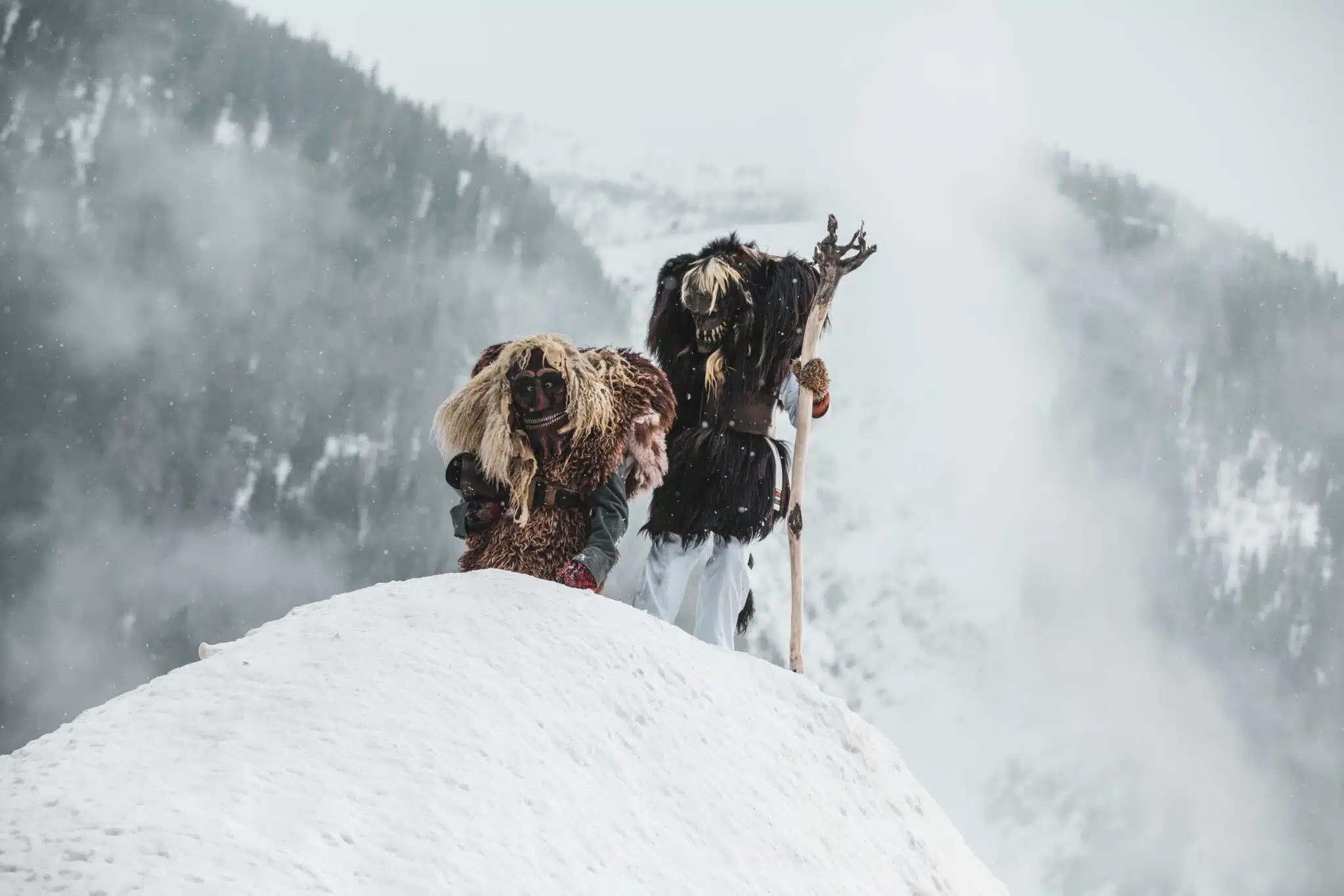 Au loin, sur la crête enneigée d'une montagne, 2 personnages avec des masques traditionnels valaisans nous regardent. Ils sont vêtus  de sortes de peaux et l'un d'entre eux s'appuie sur un grand bâton.