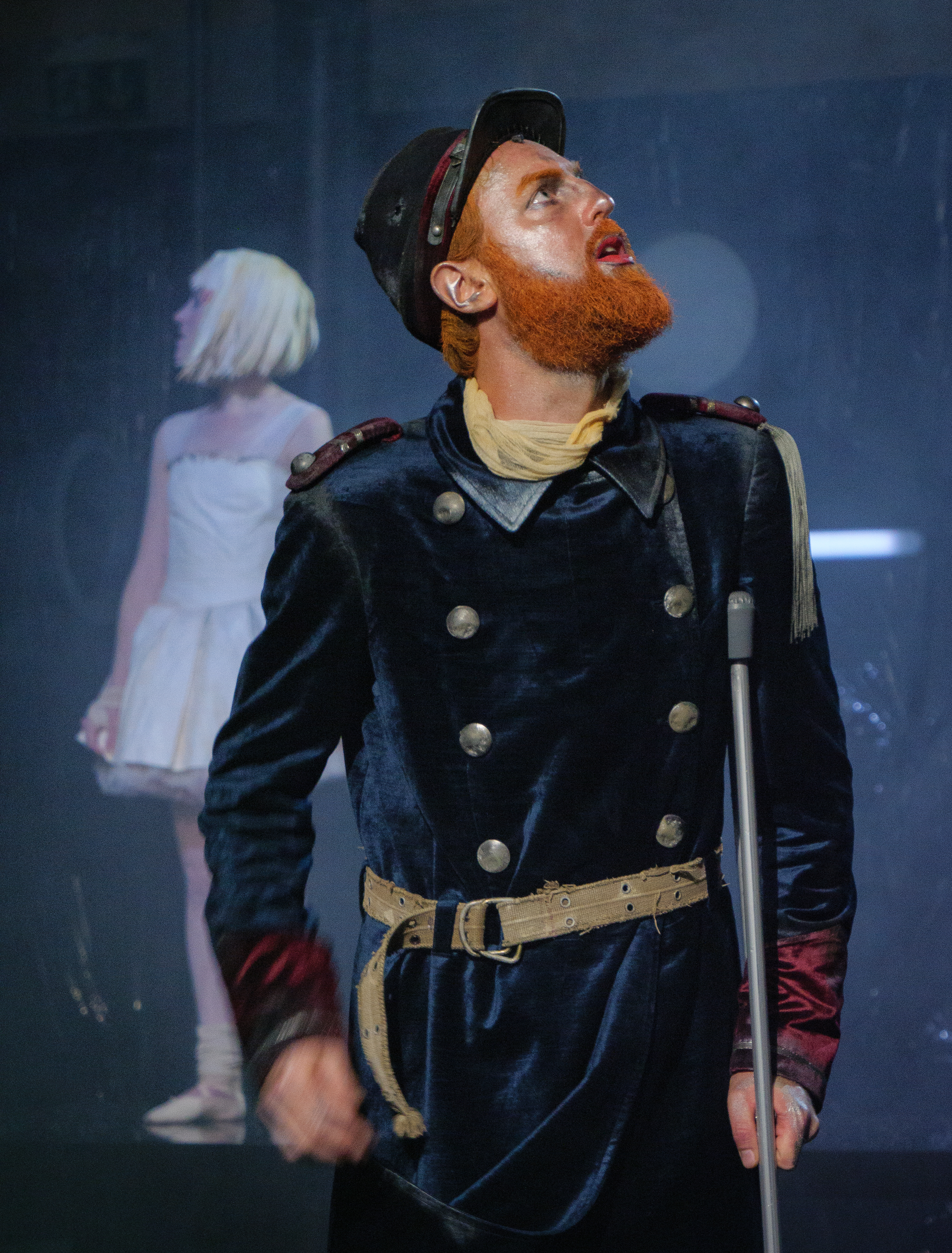Descriptif de l'image : Une comédienne vêtue d'une jupe blanche brandit un témoin lumineux orange; derrière elle se trouve un comédien vêtu d'un uniforme et d'une casquette de soldat ainsi que d'un gilet de sauvetage orange.
