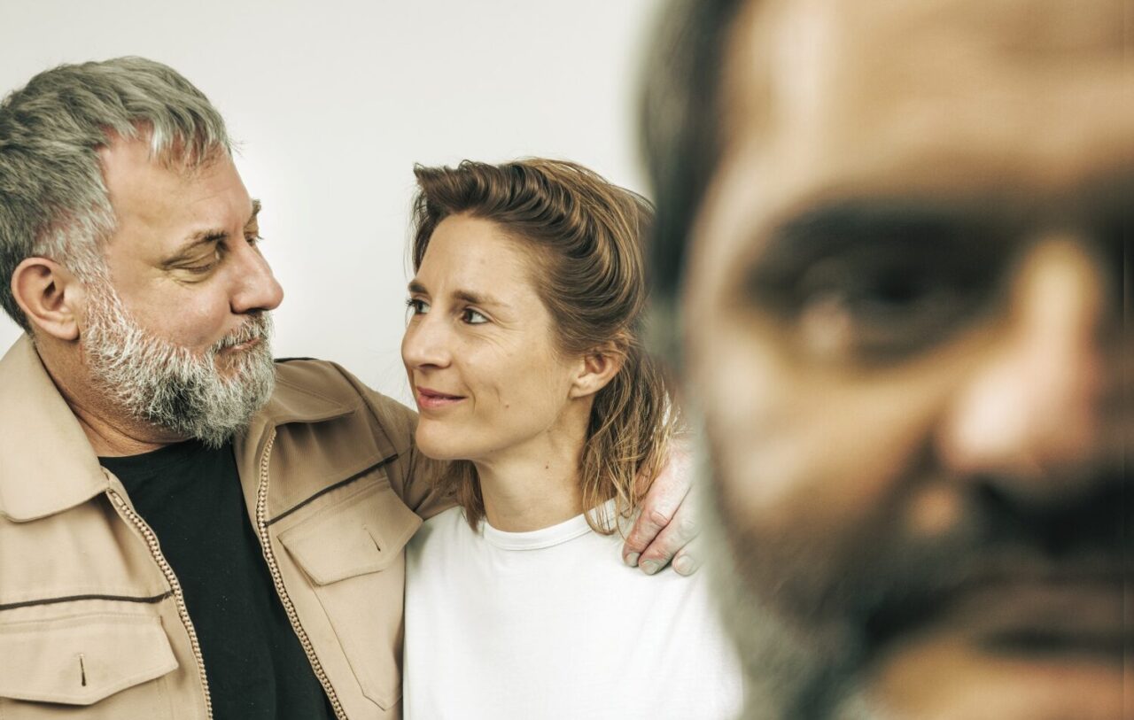 Au premier plan à droite la moitié d'un visage flouté d'un homme barbu. An arrière plan à gauche un homme barbu grisonnant regarde une jeune femme; il a la main posée sur son épaule.