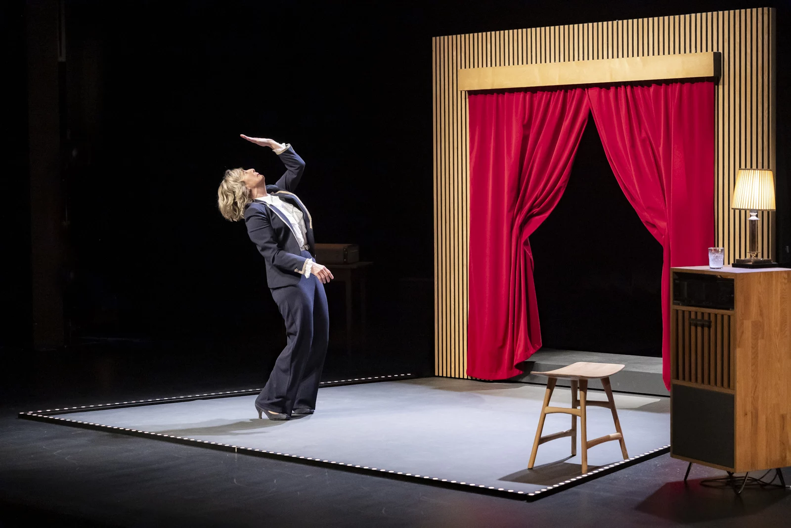 Brigitte Rosset regarde la paume de sa main en courbant le dos. A côté d'elle se trouve une mini scène avec un rideau rouge ouvert. En face d'elle, il y a un tabouret ainsi qu'un petit meuble allongé verticalement sur lequel sont posés une lampe de chevet et un verre d'eau.