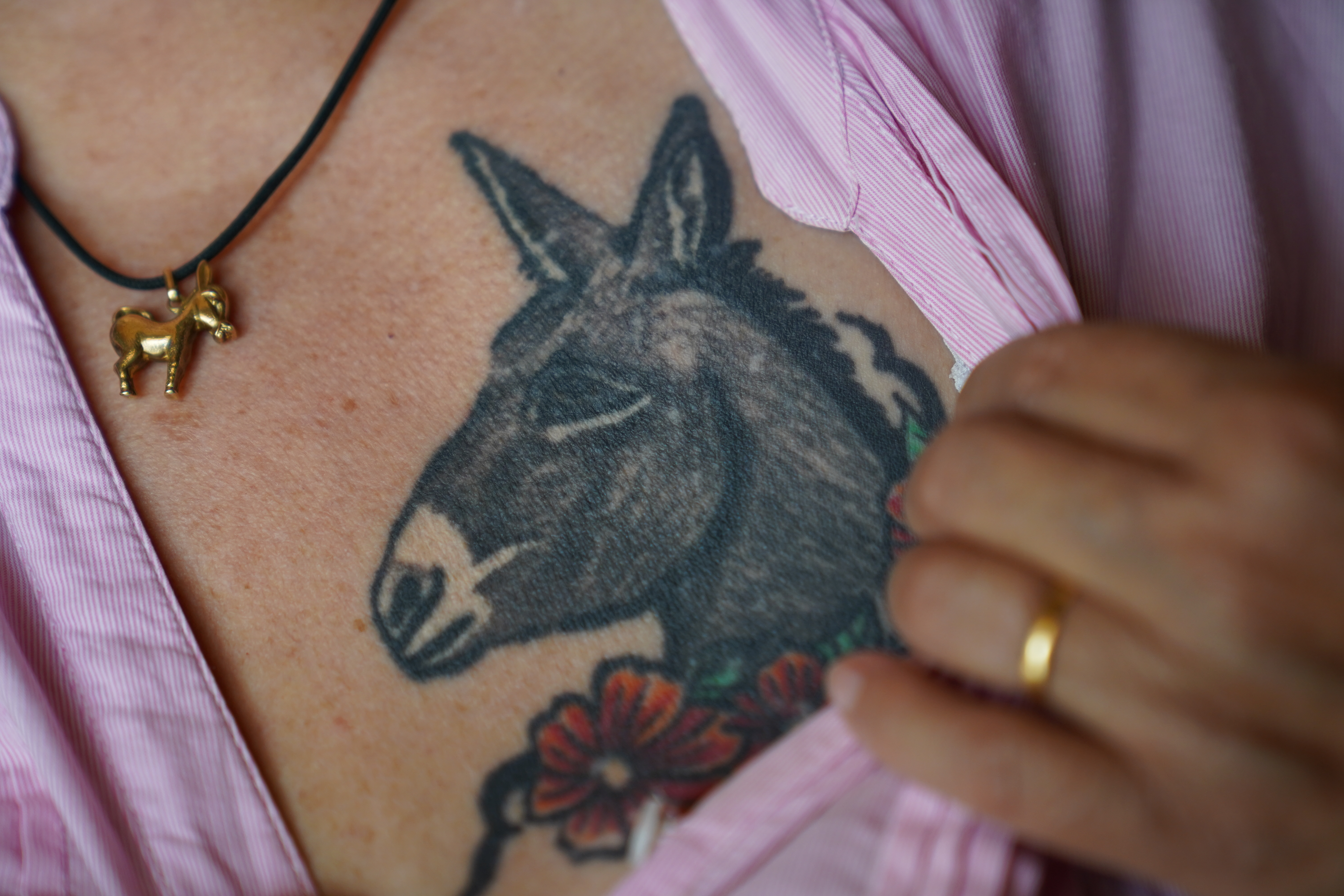 Descriptif de la photo: Détail du buste d'une femme. De sa main, elle écarte un pan de sa chemise pour dévoiler le tatouage d'une tête d'âne.