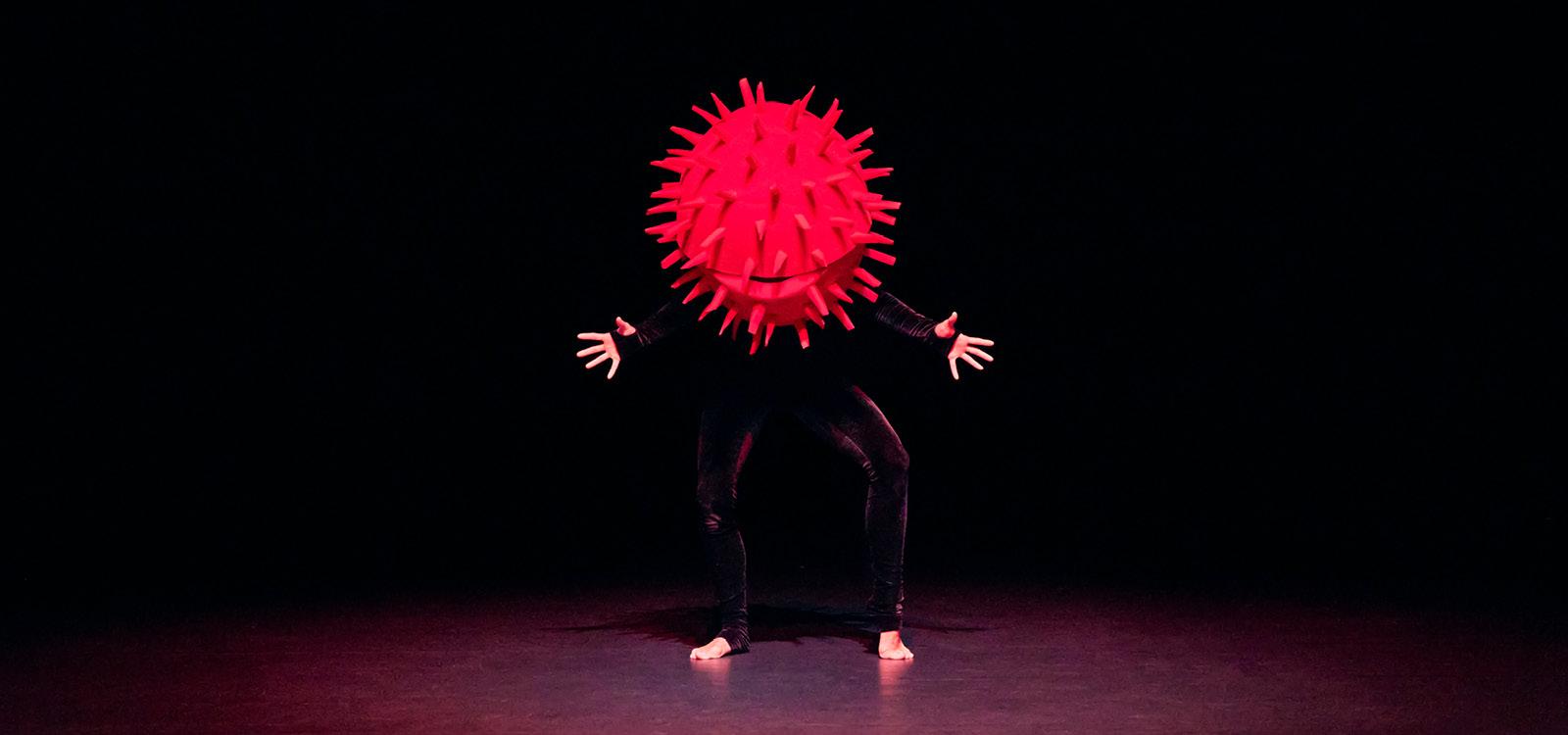 Un comédien muni d'un gros maque-tête rouge en forme de boule avec des piquants se tien face public avec les mains écartées.
