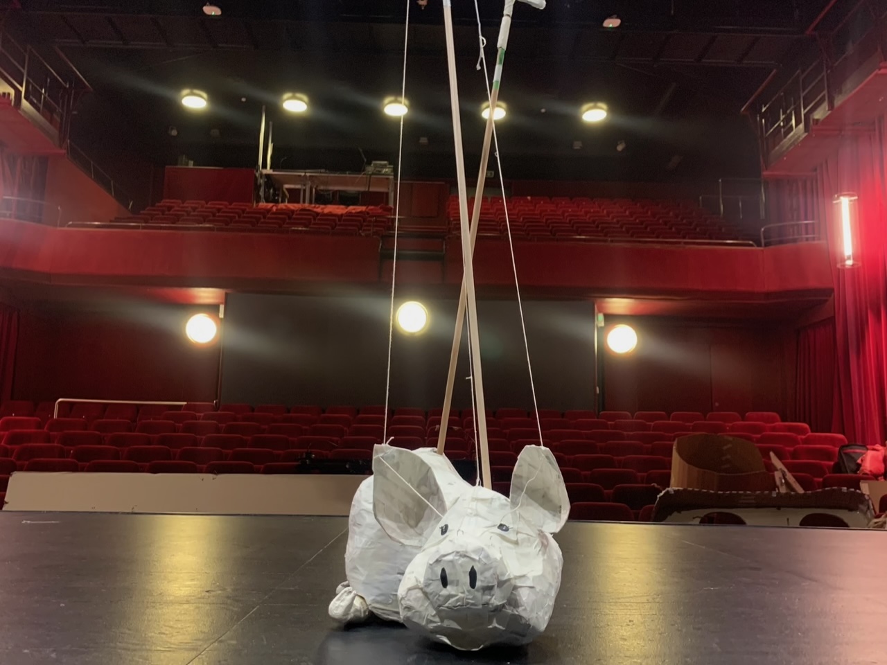 Devant les gradins rouges d'un théâtre, la marionnette d'un cochon en papier est posée sur la scène.
