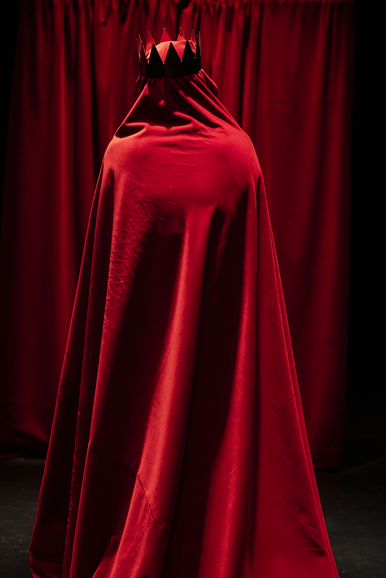 Devant un rideau rouge, se tient de dos un personnage entièrement recouvert d'une cape rouge. Il porte une couronne rouge.