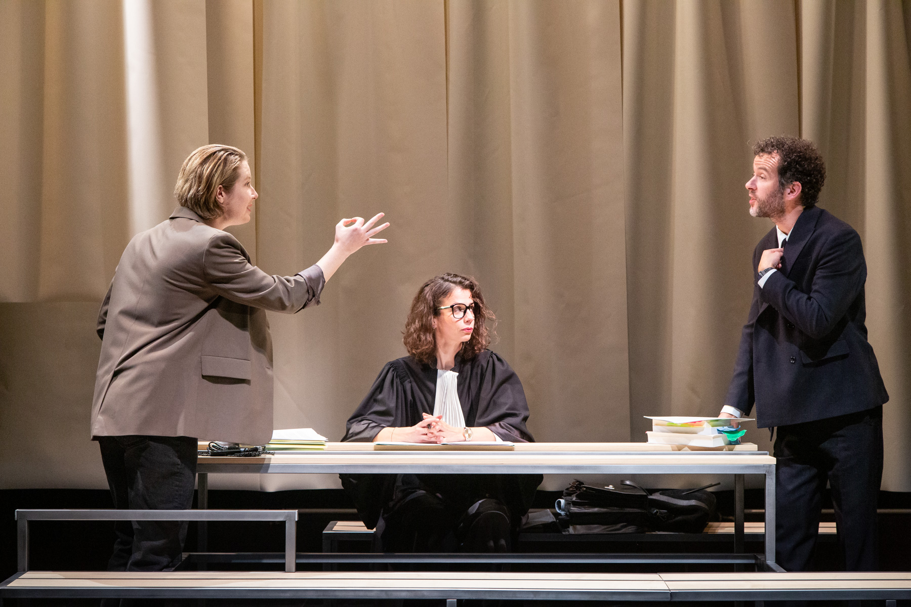 A une table de bureau est assise une femme qui porte l'habit d'un juge. De part et d'autre de la table une femme debout qui semble invectiver l'homme qui lui fait face et qui prend un air innocent en se pointant du doigt.