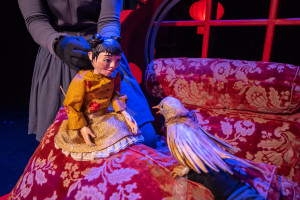 A gauche de l'image Lynn, petite marionnette aux cheveux foncés remontés en 2 chignons porte une robe jaune; elle fait face à un rossignol. Tous deux sont installés sur en canapé de couleur rouge dans le palais de l'Empereur de Chine